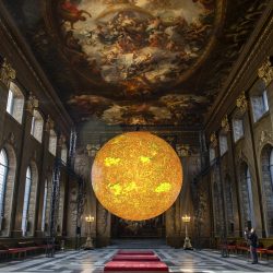 A large orange installation, Helios, which represents the sun, suspended in the middle of the Baroque style Painted Hall at the Old Royal Naval College.