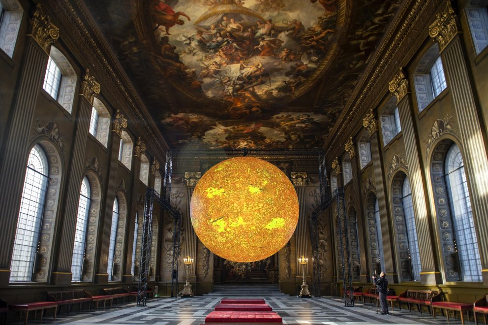 A large orange installation, Helios, which represents the sun, suspended in the middle of the Baroque style Painted Hall at the Old Royal Naval College.