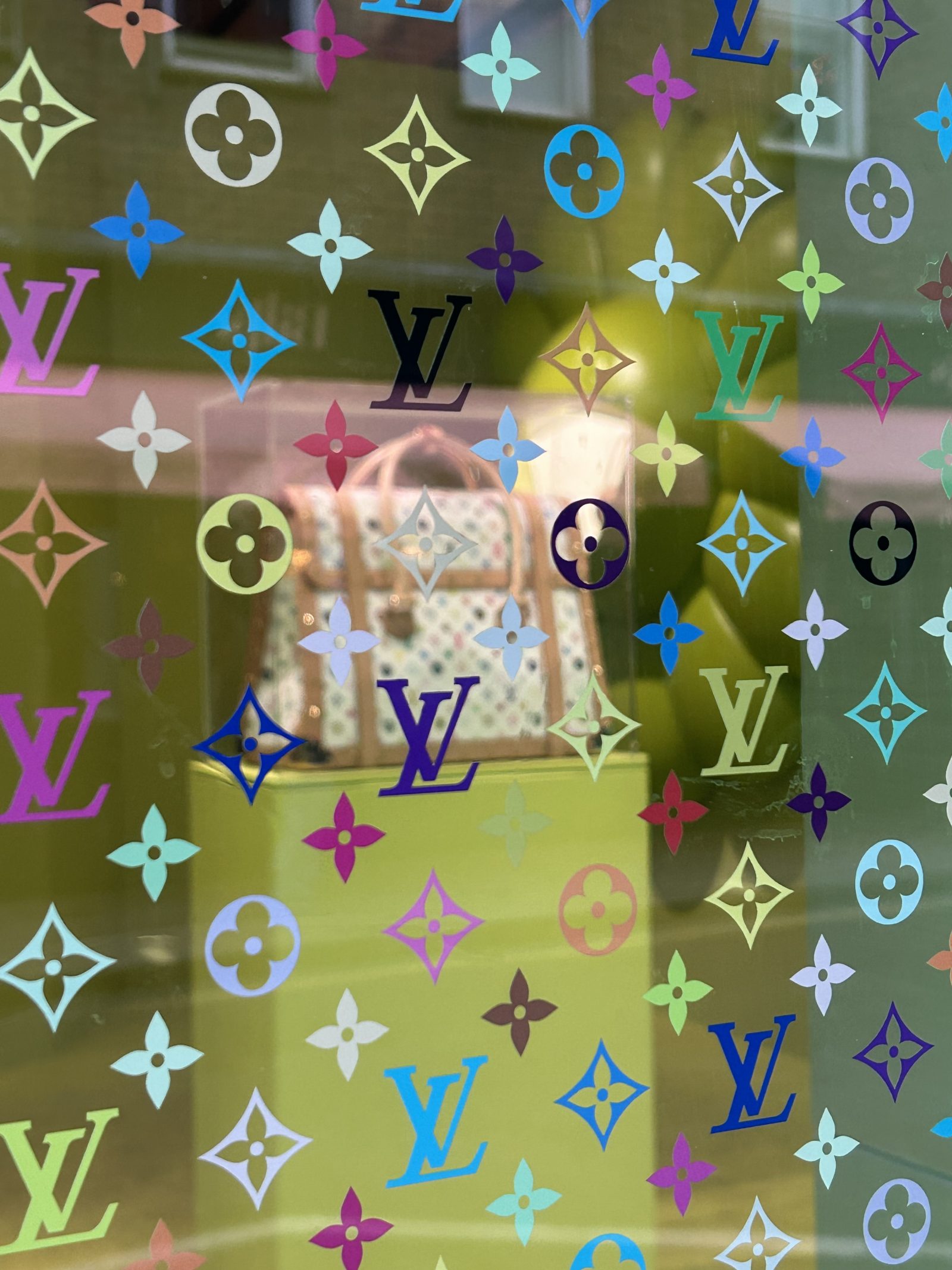 A view inside the window of the Louis Vuitton x Murakami pop-up shop in Brewer Street, London, displaying a handbag and bold monogrammed window artwork.