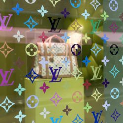 A view inside the window of the Louis Vuitton x Murakami pop-up shop in Brewer Street, London, displaying a handbag and bold monogrammed window artwork.