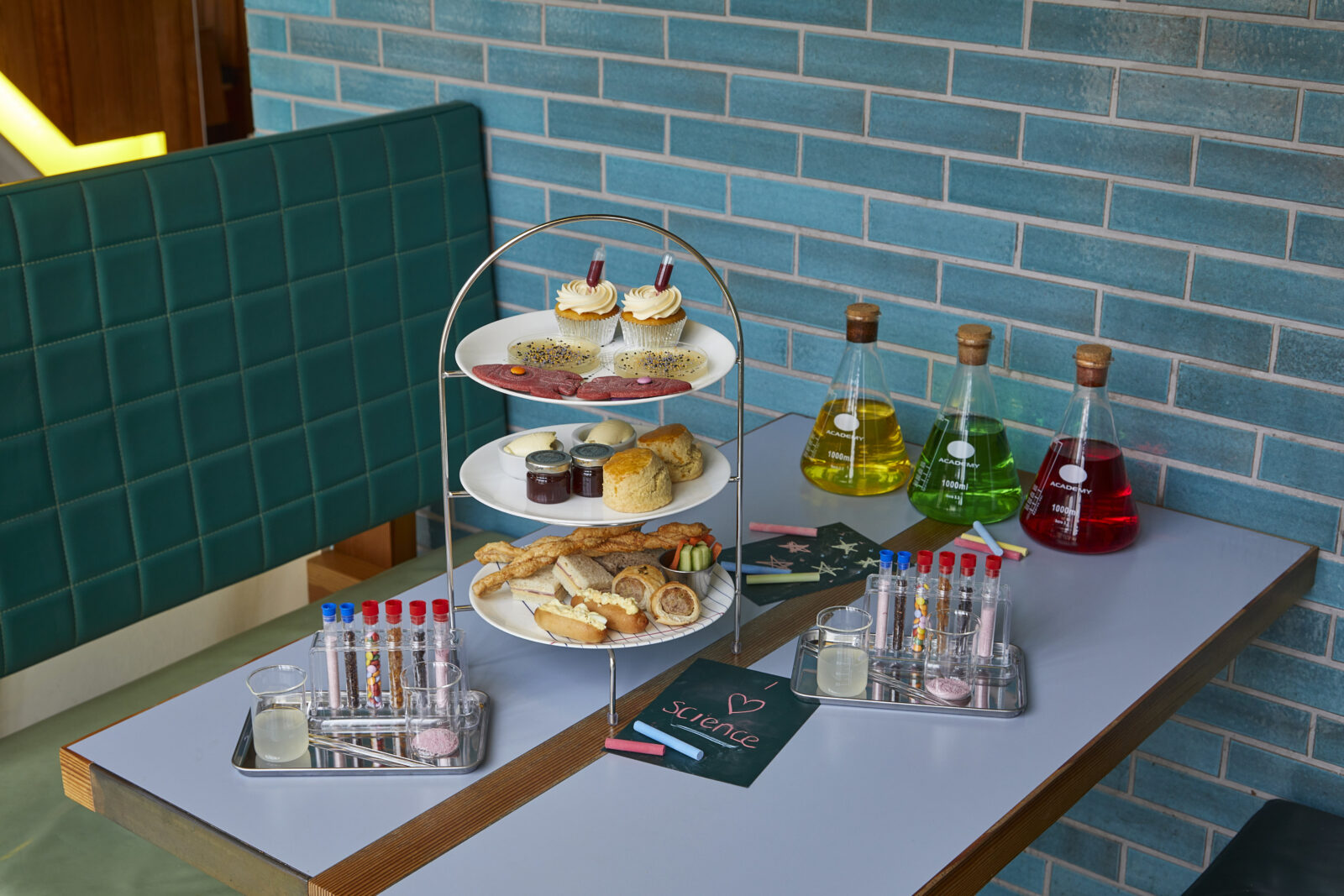 A selection of sandwiches, a tiered platter of cakes and cookies, and beakers and test tubes of coloured liquids and toppings all set out in a table.