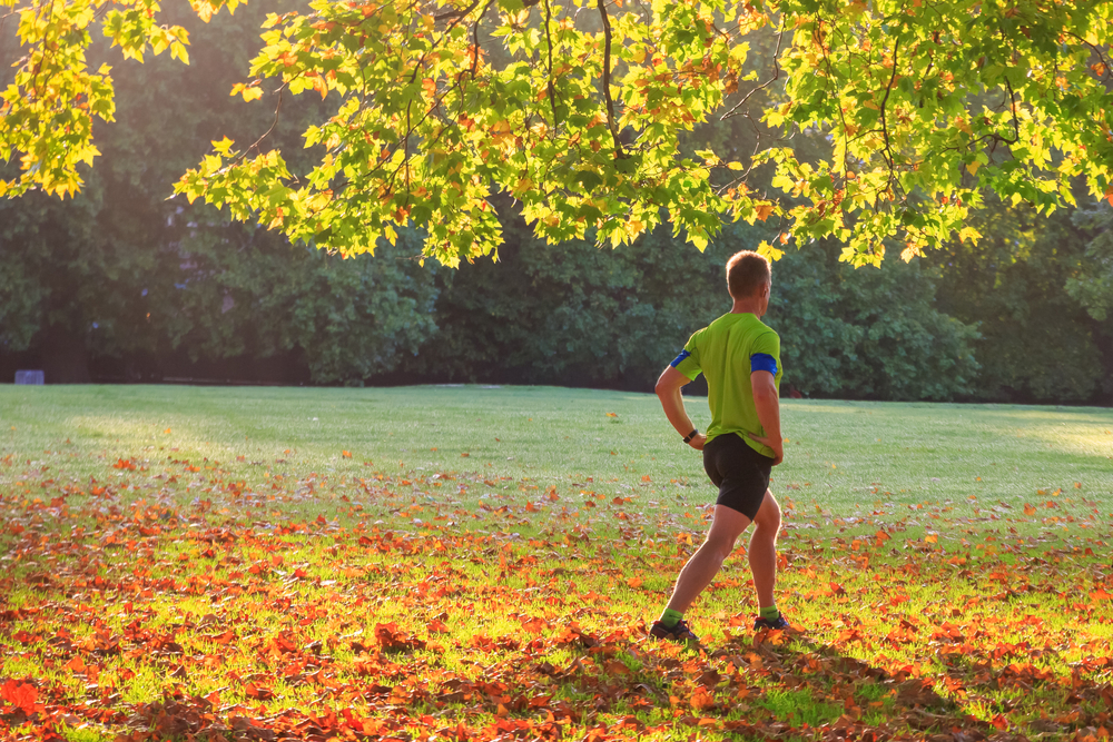 A man in shorts and tshirt, doing warm up exercises in a park on an autumnal day.