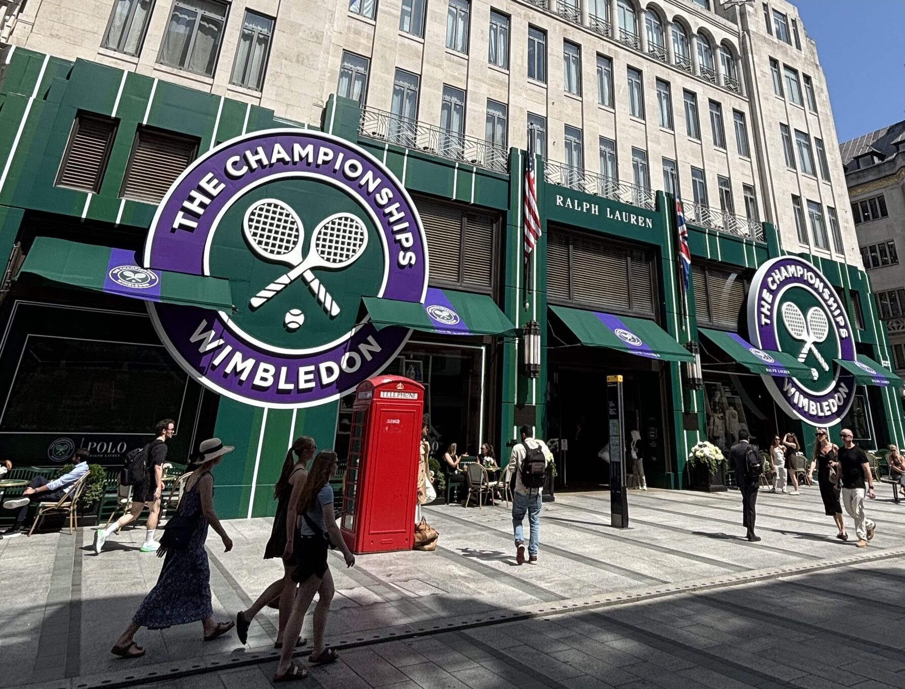 Exterior of Wimbledon shopfronts with large circular Wimbledon logo and crossed tennis rackets; pedestrians walk by with a red British telephone booth nearby.