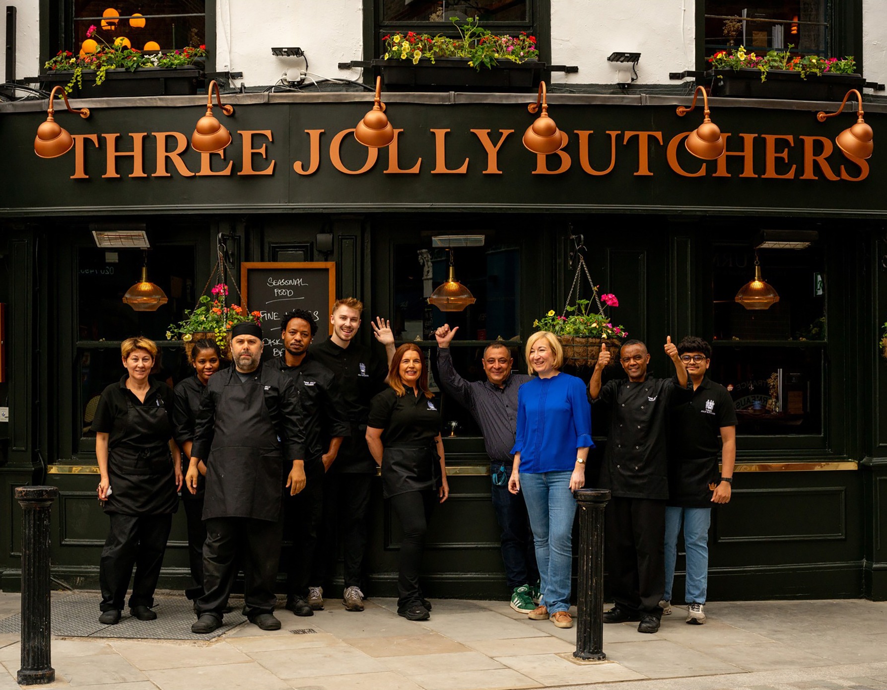 Staff group posing outside the Three Jolly Butchers restaurant in black uniforms; one person wears a bright blue top and smiles at the camera.