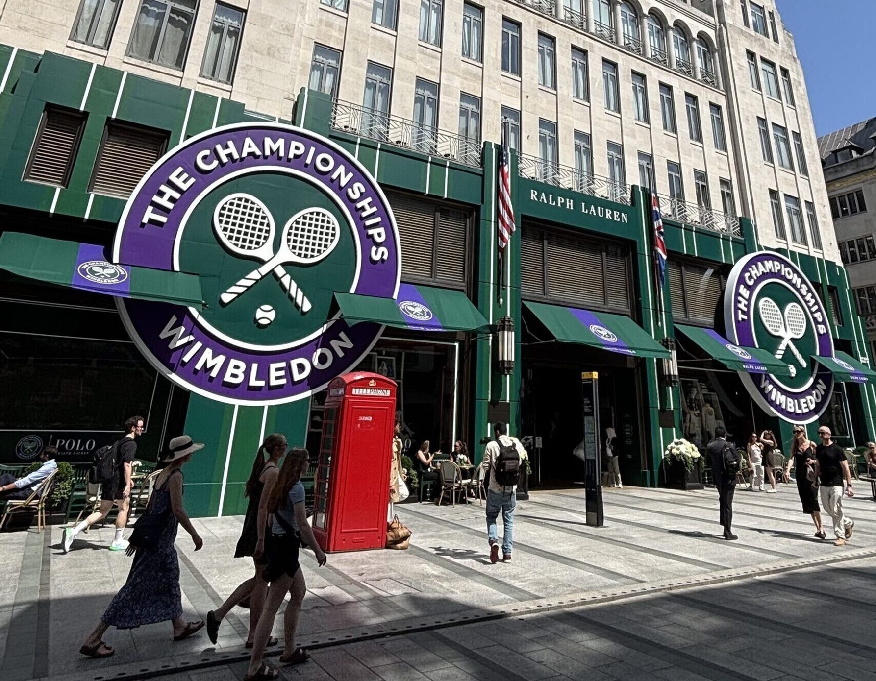 Exterior of Wimbledon shopfronts with large circular Wimbledon logo and crossed tennis rackets; pedestrians walk by with a red British telephone booth nearby.