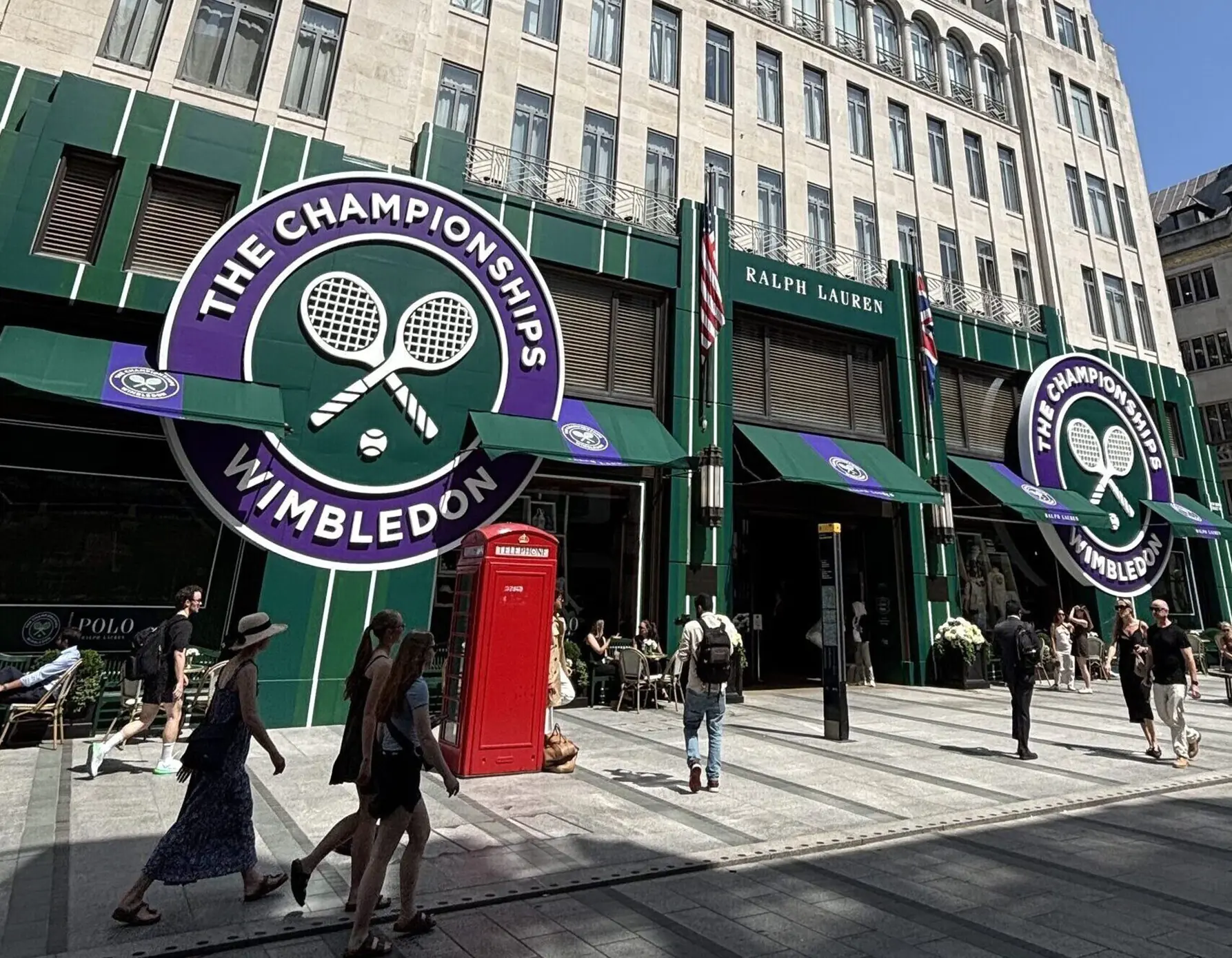 Exterior of Wimbledon shopfronts with large circular Wimbledon logo and crossed tennis rackets; pedestrians walk by with a red British telephone booth nearby.