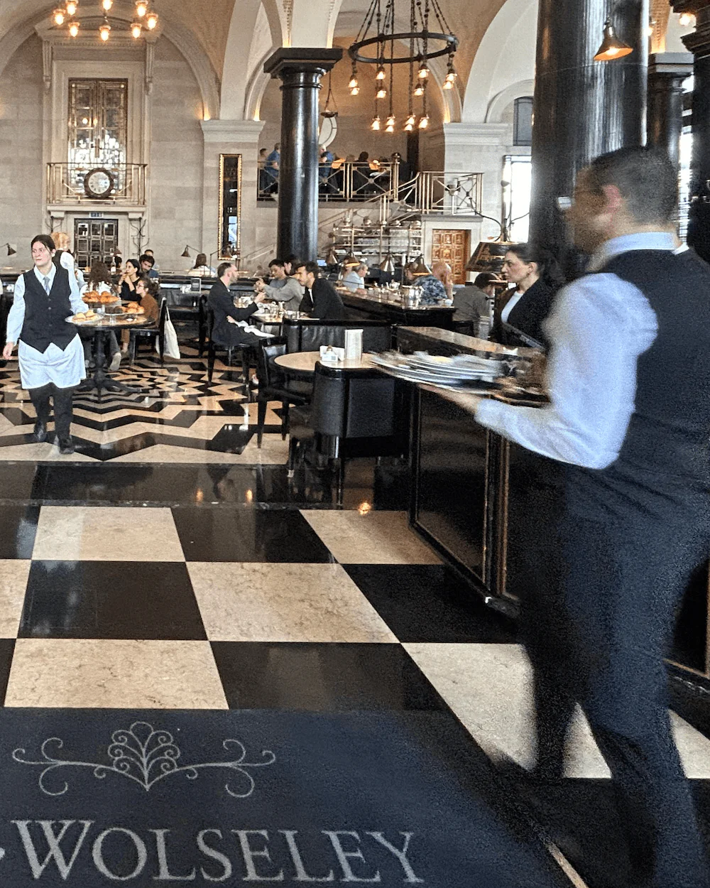 Elegant hotel dining hall with a black-and-white checkered floor, chandeliers, and arched ceilings; staff carry trays as diners sit at tables and a decorative floor mat reading 'WOLSELEY' is in the foreground.